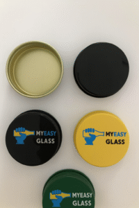 Four round 42mm aluminium caps in metallic gold, glossy black, matte yellow, and deep green, featuring MYEASY GLASS printed logo, suitable for spice jar packaging
