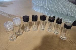 Seven clear round glass spice jars with various functional lids including transparent grinders, black plastic shaker tops, dual-flap lids, and pour holes, arranged in front of bulk inventory.