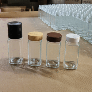 Four empty square glass spice jars with assorted lids including a black grinder cap, bamboo cap, dark wood cap, and a white plastic shaker cap, displayed on a factory tabletop.