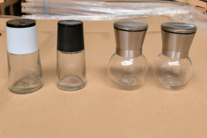 Four empty glass spice grinder bottles shown with different lid types including black and white plastic grinder tops and stainless steel lids, ideal for whole pepper or salt packaging.